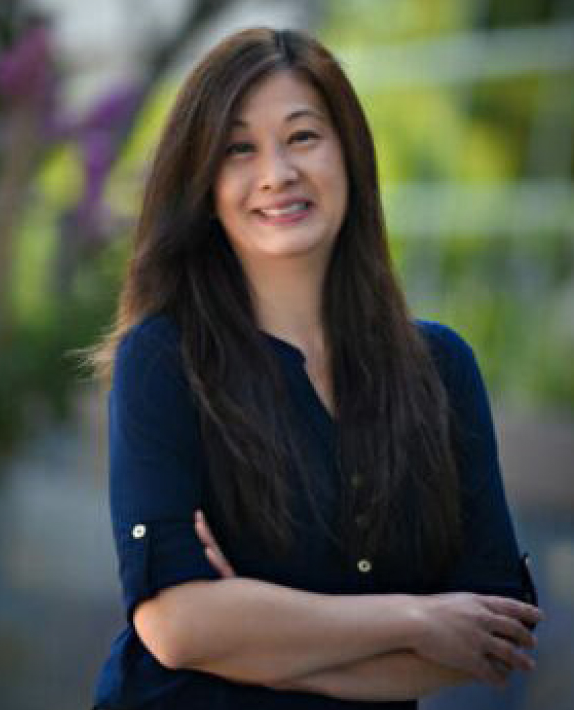 Smiling woman with long dark hair wearing a navy blue shirt, arms crossed, outdoors with a green blurred background.