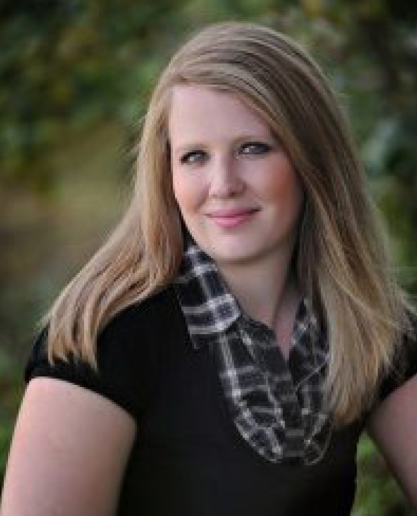 Portrait of a woman with long blonde hair, wearing a black top and plaid scarf, outdoors with greenery in the background.
