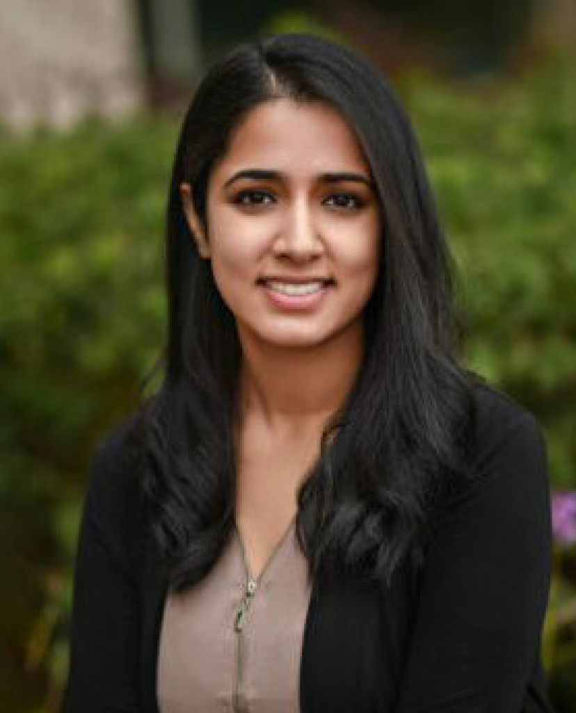 Portrait of a young woman with long black hair smiling outdoors, wearing a beige top and black cardigan, with greenery in the background.