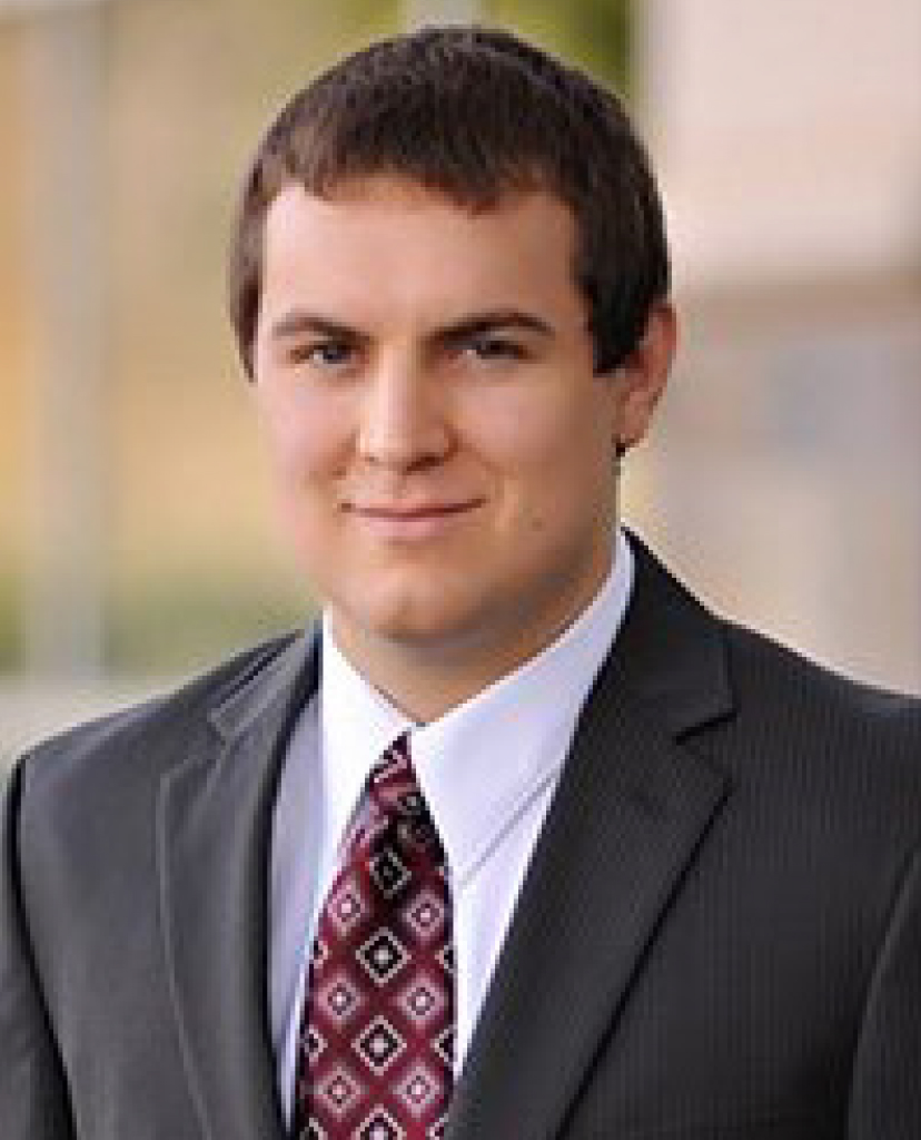 Professional headshot of a man in a dark suit and red patterned tie, smiling slightly in a neutral background.