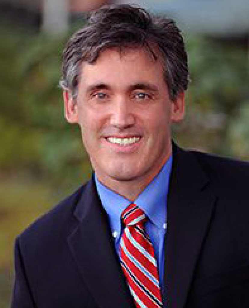 Smiling man in a dark suit with a blue shirt and red striped tie, outdoors.